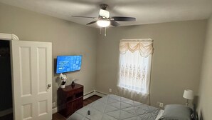 Une chambre à coucher comprenant un lit, un ventilateur de plafond, une télévision, une fenêtre avec des rideaux et une porte.