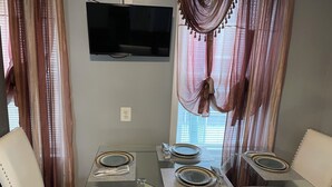Un coin repas comprenant une table en verre, deux chaises blanches rembourrées et un téléviseur à écran plat fixé au mur.