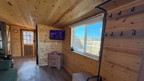 Interior de una cabaña de madera con piso de madera, un televisor montado en la pared, una ventana que ofrece vistas al exterior y un banco junto a la ventana.