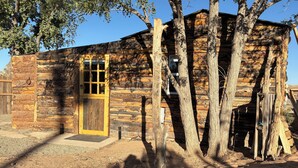 Una cabaña rústica de madera con una sola puerta y ventana, rodeada de árboles y un sendero de grava.