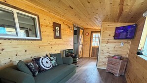 Un interior de cabaña acogedor con un sofá verde, paredes de madera, un televisor de pantalla plana y un pequeño cofre de madera.