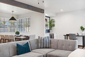 Ein modernes Wohnzimmer mit grauem Sofa, einem Essbereich mit Tisch und Stühlen, einer Küchenzeile mit Schränken und einem großen Fenster mit Jalousien.