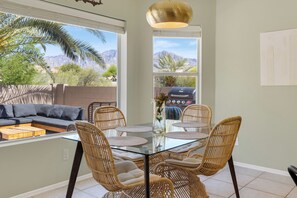 Un comedor con mesa de vidrio rodeada de sillas de mimbre, un sofá, y una parrilla visible a través de la ventana.