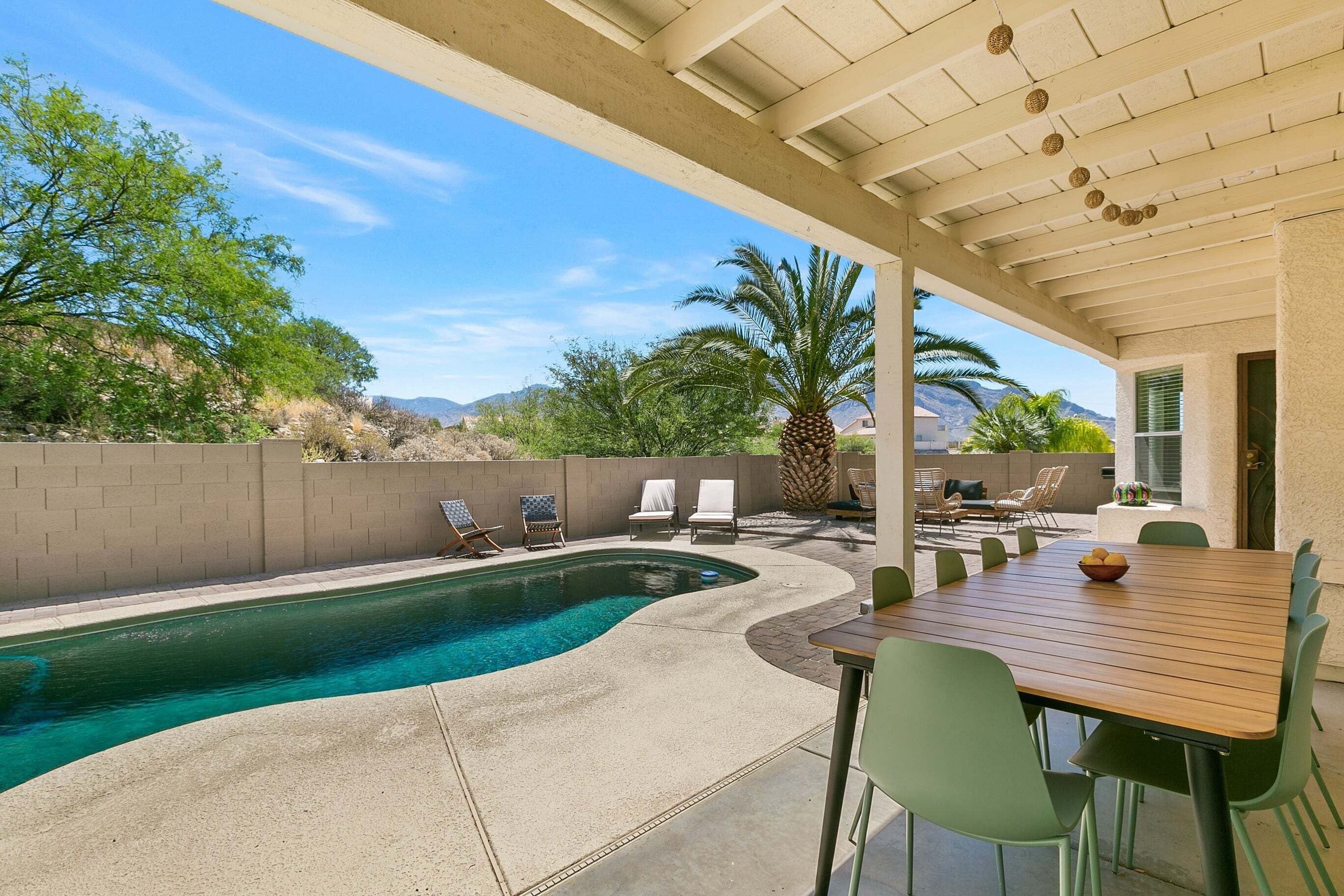 A covered outdoor area with a swimming pool, lounge chairs, a dining table, and a view of mountains.