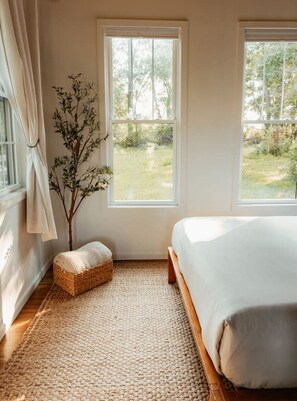 Een slaapkamer met een bed, twee ramen, een kleine plant en een geweven vloerkleed.
