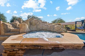 A hot tub surrounded by stone rocks, a pool, and a covered seating area.