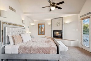A bedroom with a bed, ceiling fan, fireplace, and a window with blinds.