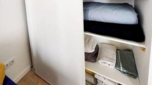 A room with a white wardrobe, a bed with a blue and black mattress, and a shelf with pillows and folded clothes. - Apparttement distrito10 (Paris)