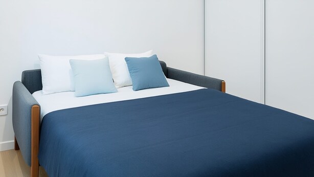 A bed with a navy blue bedspread and two pillows, one white and one light blue, in a room with a white wall and wooden floor. - Apparttement distrito10 (Paris)
