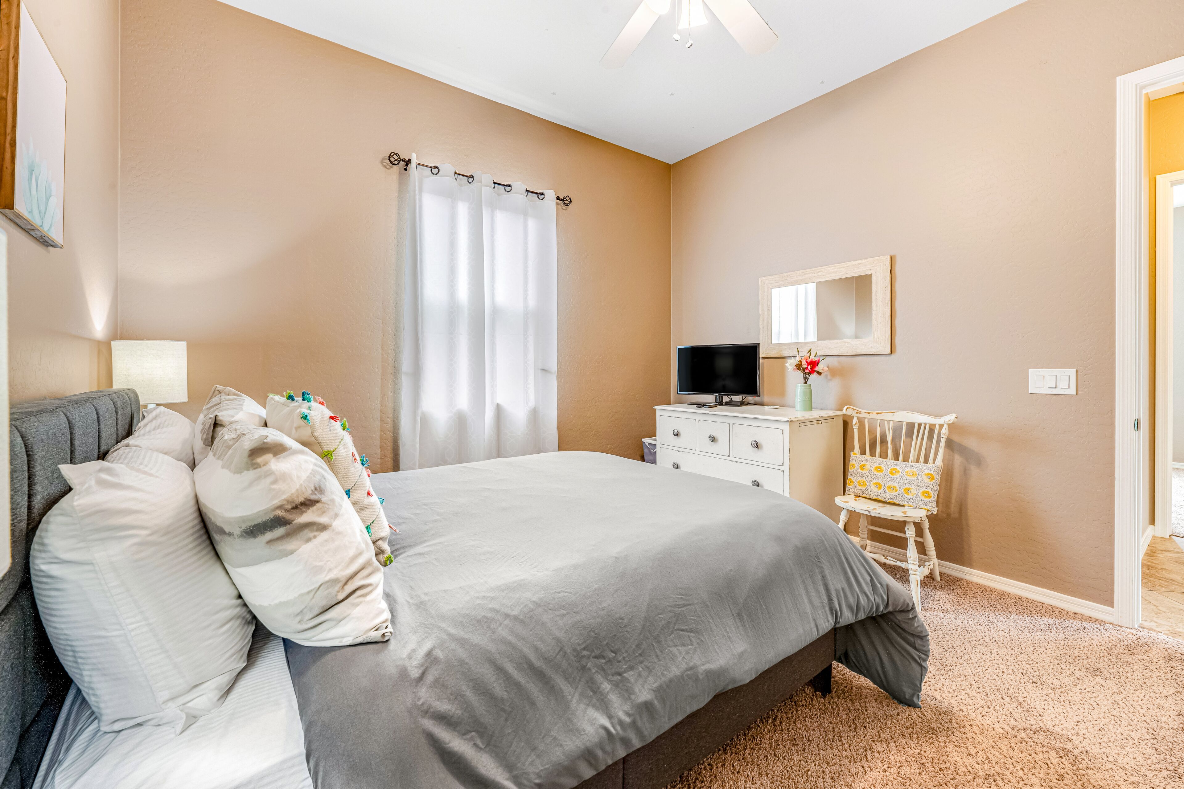 Une chambre à coucher avec un lit, une commode, une chaise, un téléviseur et une fenêtre avec des rideaux.