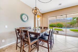 A dining area with a large table and chairs, a chandelier, a clock, and a view of a landscaped outdoor area with a pool. - South Mountain-view home with private pool & Ping-Pong - near hiking trails (Phoenix)