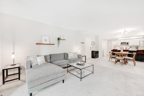 A modern living room with a sectional sofa, a glass coffee table, and a dining area with wooden chairs and a table. - Stylish updated condo with balcony, pool, & hot tub - near golf and greenway (Scottsdale)