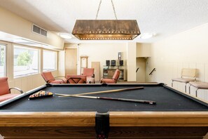 Une table de billard, avec des queues et des billes, entourée de fauteuils en cuir, dans une pièce dotée de grandes fenêtres et d’un luminaire suspendu.