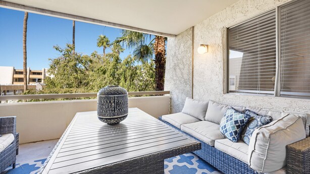 A balcony with a wicker sofa, a patterned table, and a view of palm trees and buildings. - Stylish Old Town Scottsdale condo with pool, spa, gym, & game room (Scottsdale)