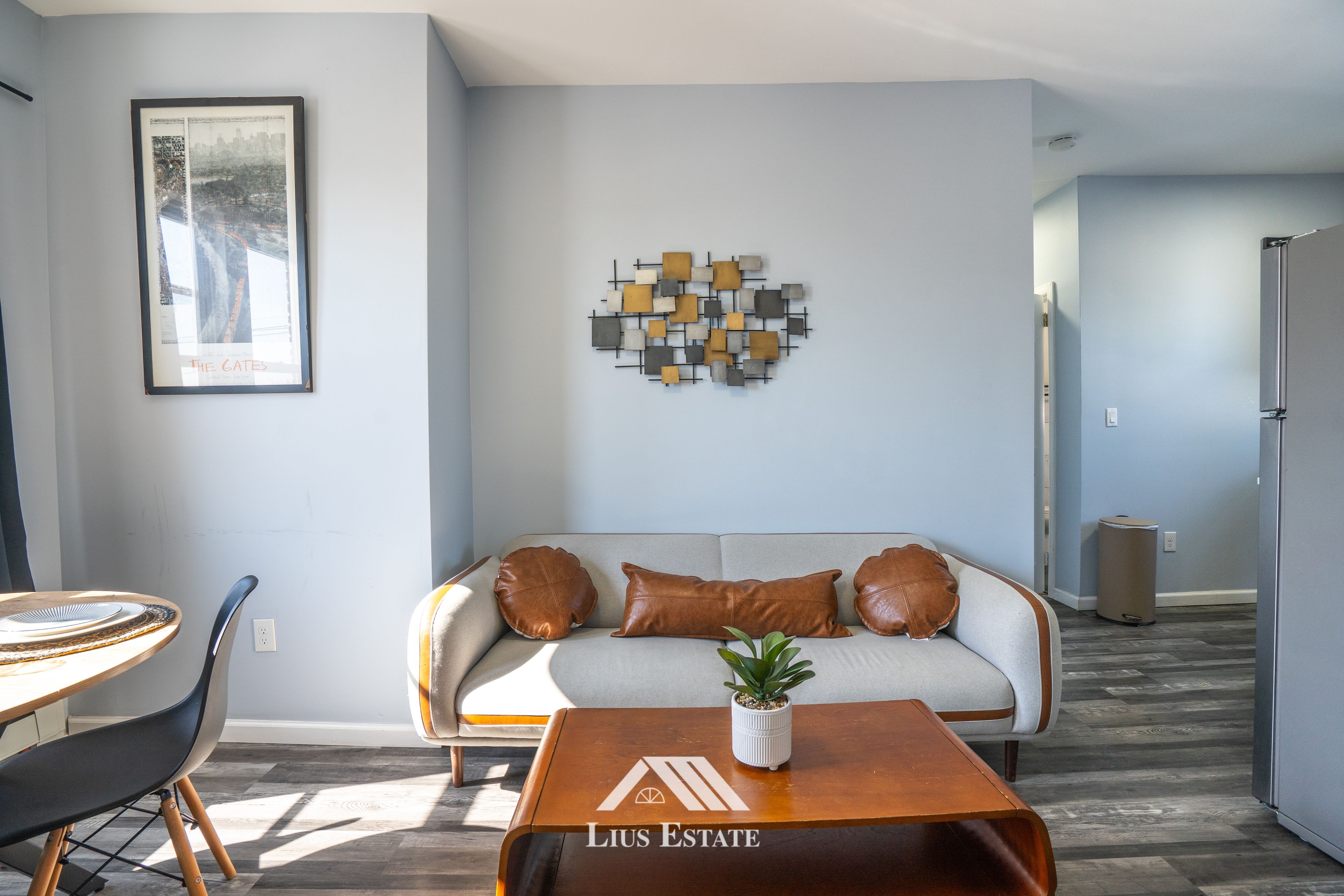 A modern living room with a grey sofa, a coffee table with a logo, a framed picture on the wall, and a hallway leading to a kitchen.