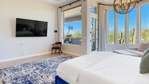 Un dormitorio con una cama grande, un televisor de pantalla plana montado en la pared, un ventilador de techo y vistas al exterior a través de amplias ventanas.