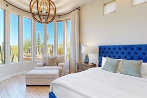 A bedroom with a large bed, a sofa, a nightstand, and a view of cacti outside.