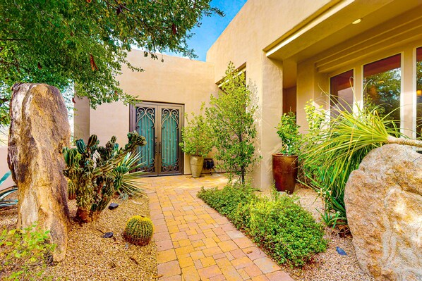 A pathway leading to a building with a decorative metal gate, surrounded by various plants and large rocks.