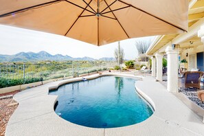 A swimming pool with a curved design, surrounded by a patio area with outdoor furniture and a large umbrella providing shade. - Mountain-view home with pool, furnished patio, washer/dryer & central AC (Fountain Hills)