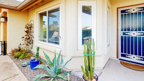 Una casa con fachada blanca, una puerta azul con detalles de metal decorativo y un pequeño jardín con diversas plantas y un cactus.