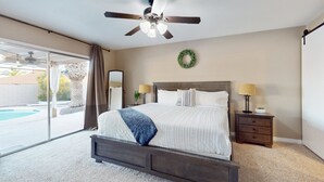 A bedroom with a large bed, a ceiling fan, a nightstand with lamps, a wall-mounted mirror, and a view of a swimming pool through a sliding glass door. - Charming, dog-friendly home only minutes to Old Town with pool & custom bar (Scottsdale)