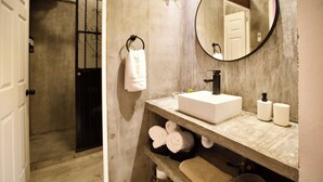 Une salle de bain avec un lavabo blanc, un miroir rond et un porte-serviettes sur lequel est posée une serviette blanche. On peut voir deux bouteilles sur le comptoir du lavabo.