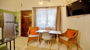 Un espace de vie compact comprenant une table à manger, deux chaises en osier, un canapé, un téléviseur et un réfrigérateur.