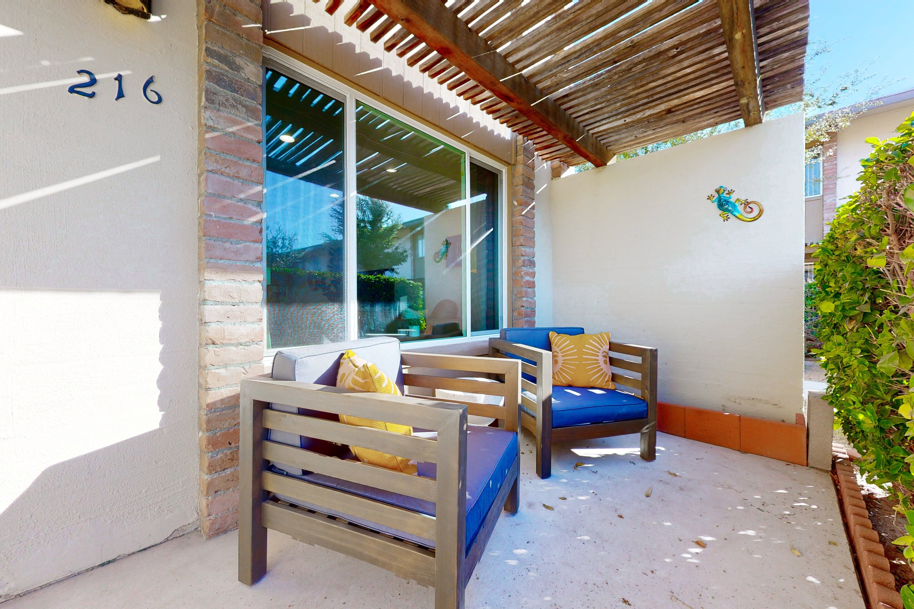 Un espace extérieur abrité, aménagé avec deux chaises en bois et un coussin bleu, un mur en briques et une fenêtre donnant sur un paysage verdoyant.