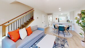 A modern living room with a grey sofa, orange and white cushions, a wooden staircase, and a dining area with a round table and chairs. - 2-story dog-friendly condo with patio, outdoor pool, W/D, streaming, partial AC (Scottsdale)