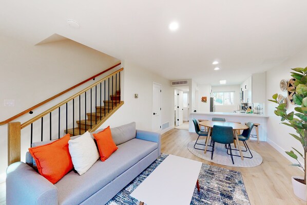A modern living room with a grey sofa, orange and white cushions, a wooden staircase, and a dining area with a round table and chairs. - 2-story dog-friendly condo with patio, outdoor pool, W/D, streaming, partial AC (Scottsdale)