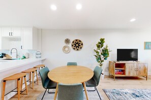 A modern kitchen with white cabinets, a wooden dining table with four chairs, a TV on a wooden cabinet, and decorative wall art. - 2-story dog-friendly condo with patio, outdoor pool, W/D, streaming, partial AC (Scottsdale)