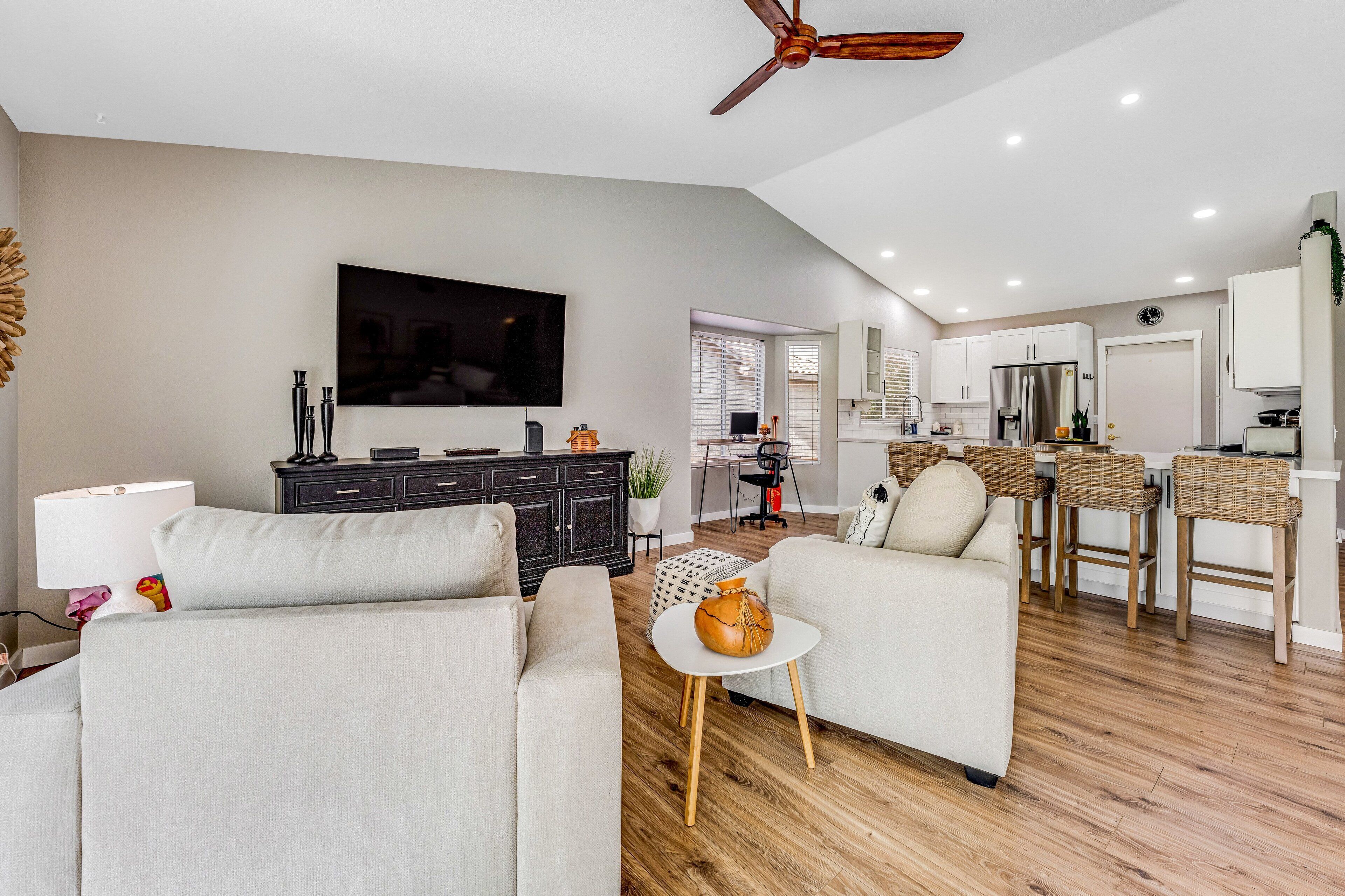 A modern living room with a sofa, a coffee table, and a TV. There is a kitchen area with a refrigerator and a dining table.