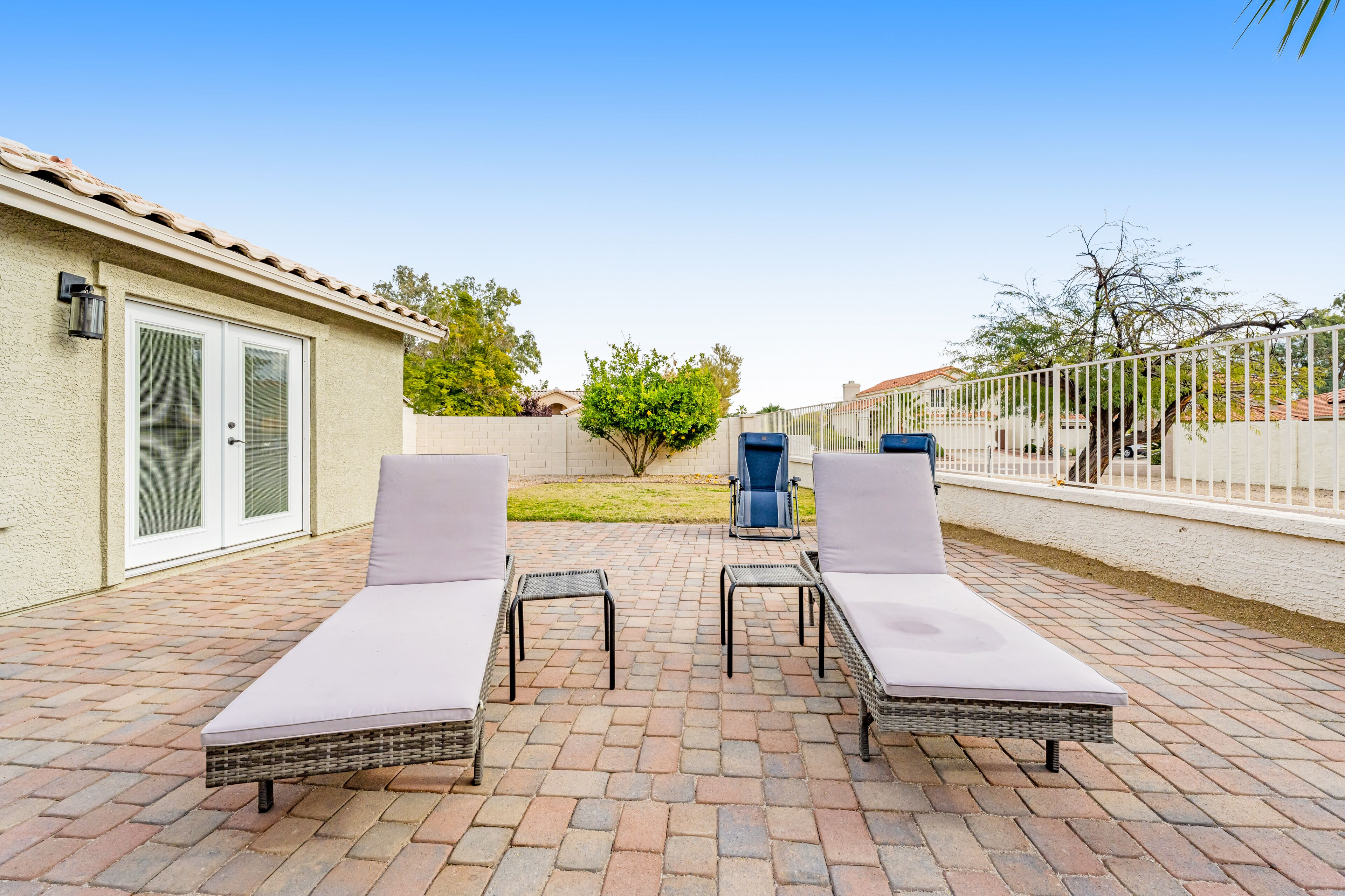 A patio with two lounge chairs and a small table, a white fence, and a tree in the background.