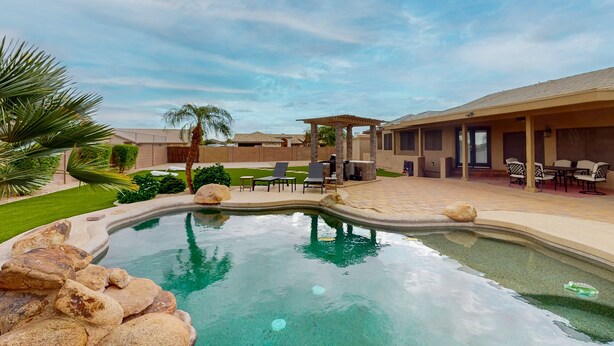 A swimming pool with a curved edge, surrounded by rocks and a patio area with outdoor furniture. - Peoria Paradise: Poolside Vibes & Family Good Times. Spacious Backyard with Pool (Peoria)