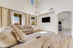 A spacious living room with a large sofa, a television mounted on the wall, and a chandelier. - Mountain-view gem - 2 large living rooms, heated pool, enclosed yard, AC & W/D (Avondale)