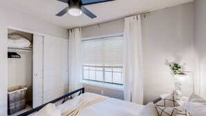 Un dormitorio con cama, ventilador de techo, ventana con persianas, mesita de noche con lámpara y armario.