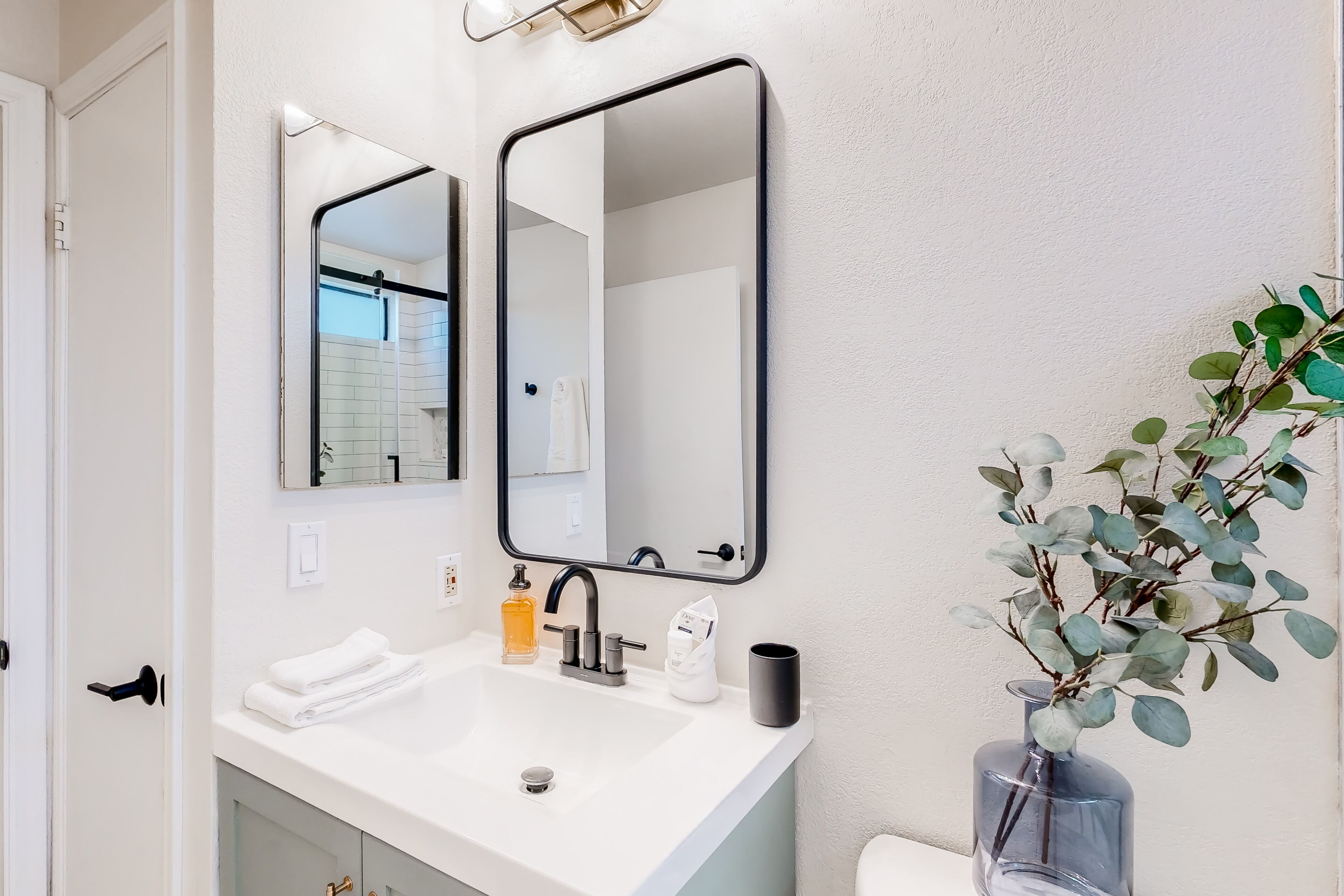 A bathroom with a white sink, two mirrors, a towel rack, a bottle of soap, a cup, and a vase with green leaves.
