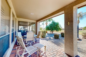 A covered patio with a table and chairs, a brick floor, and a view of palm trees and greenery. - Palm-lined home with pool access, grill, office & golf course views (Sun Lakes)