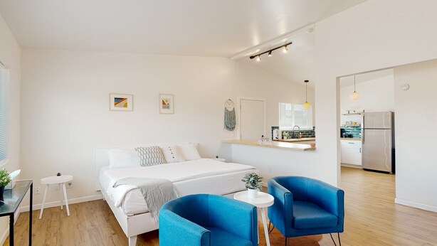 A modern bedroom with a bed, two blue chairs, a small white table, and a kitchen area visible through an open door. - Inviting dog-friendly studio with full kitchen and central AC (Tucson)
