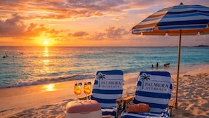Deux chaises de plage portant la marque « Palmera Retreats », un parasol et une glacière avec la marque « IceMaster », le tout installé sur une plage de sable au coucher du soleil.