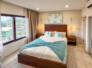 Une chambre à coucher avec un grand lit, deux tables de chevet, une fenêtre avec des rideaux, une œuvre d’art accrochée au mur et un tapis à motifs.