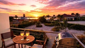 Un balcon avec une table et des chaises, un barbecue et une vue sur le coucher de soleil au-dessus d’une plage et de palmiers.