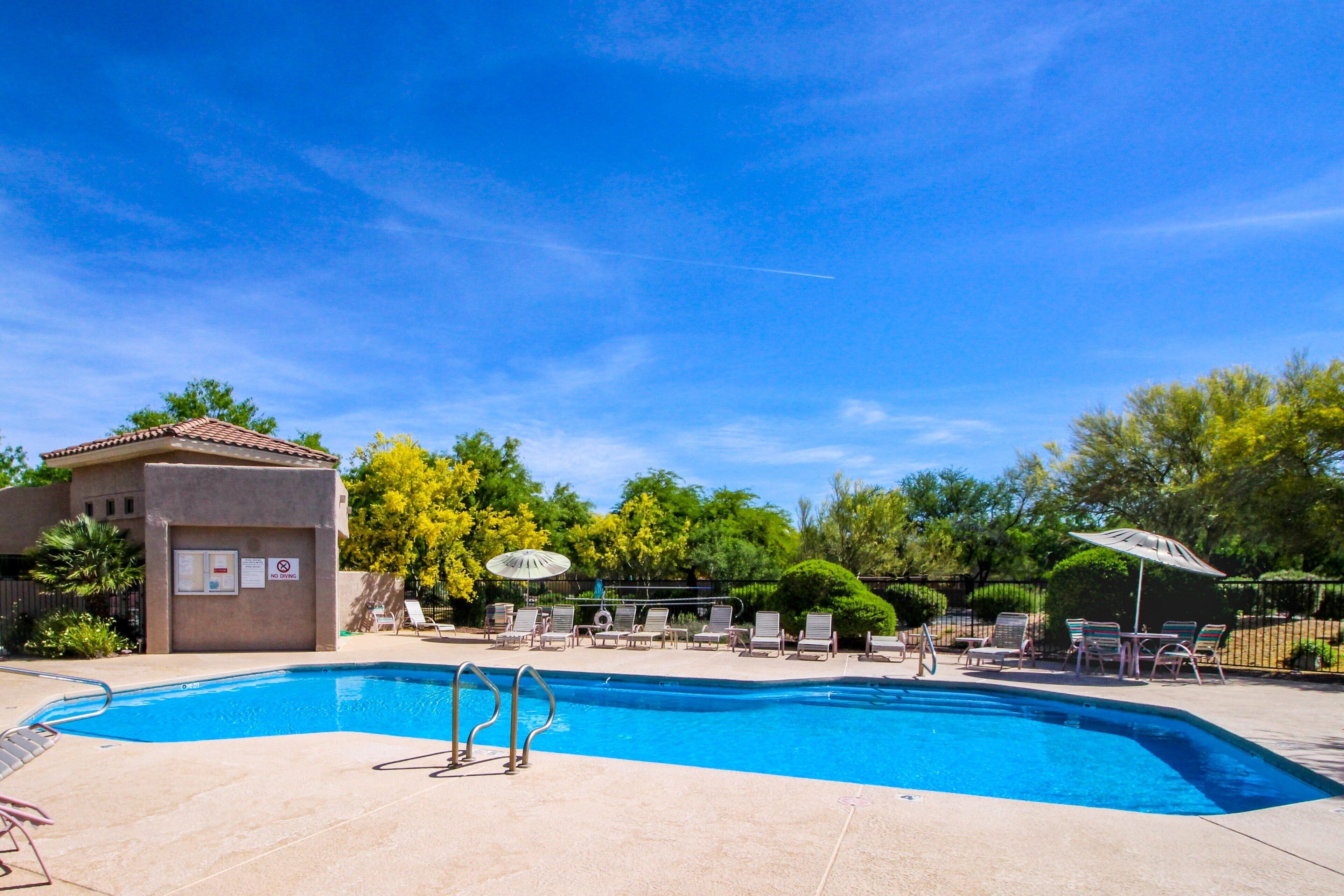 A swimming pool with lounge chairs and umbrellas, surrounded by trees and a building with a tiled roof.