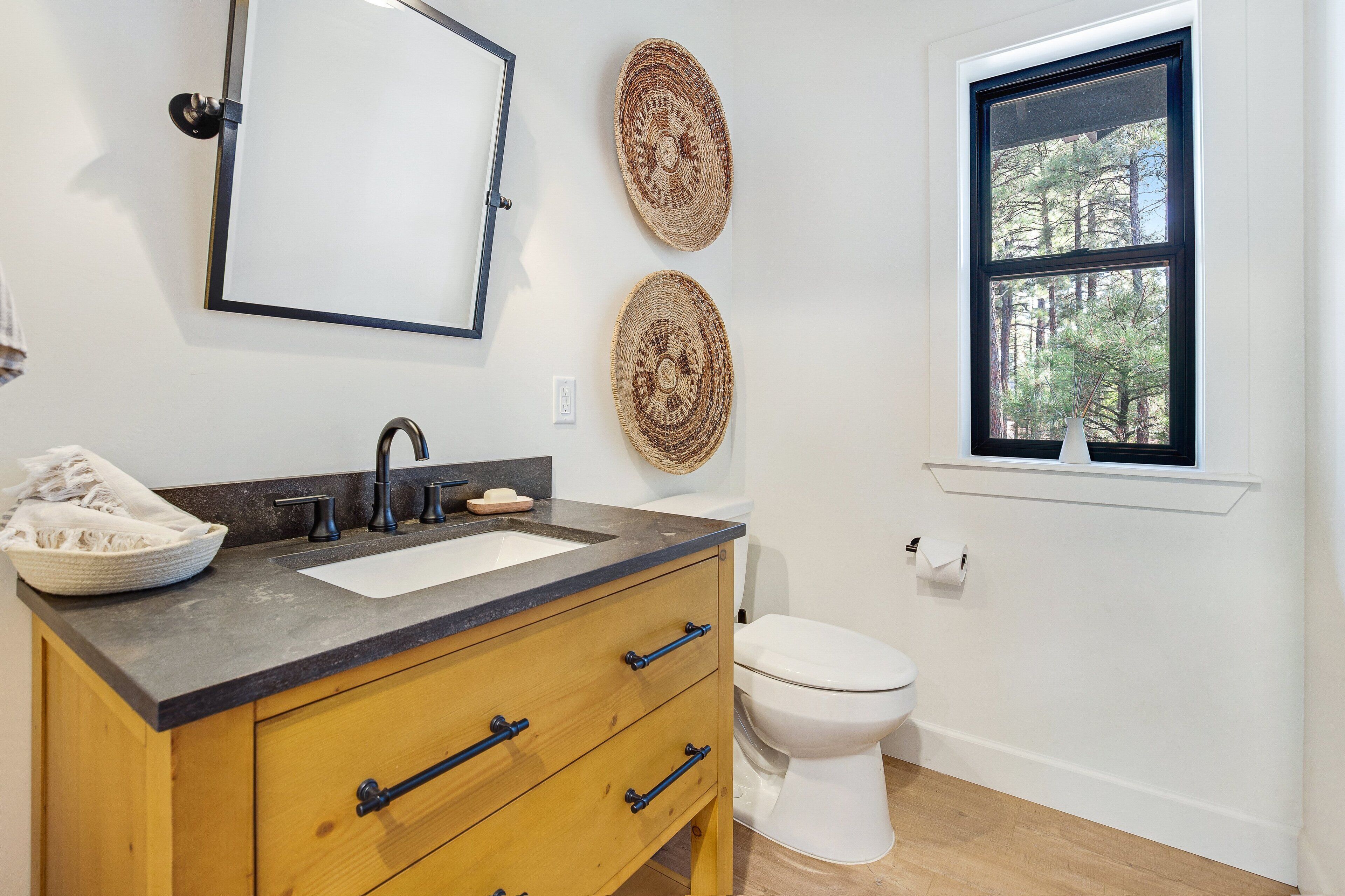 A bathroom with a wooden vanity, two woven wall decorations, a white toilet, and a window with a view of trees.