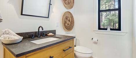 A bathroom with a wooden vanity, two woven wall decorations, a white toilet, and a window with a view of trees.