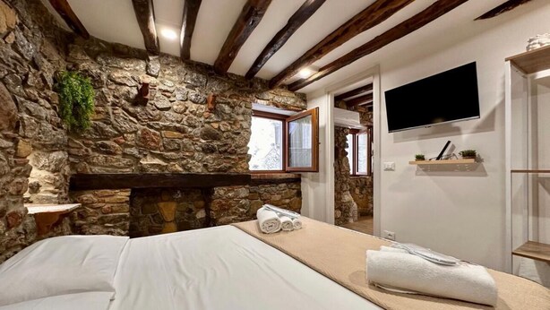 Une chambre avec un mur en pierre, des poutres apparentes, un lit avec du linge de maison blanc, un téléviseur fixé au mur et une étagère avec des objets décoratifs.