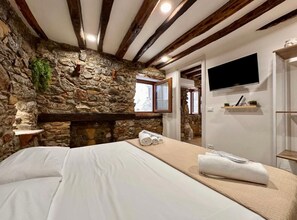 Une chambre avec un mur en pierre, des poutres apparentes, un lit avec du linge de maison blanc, un téléviseur fixé au mur et une étagère avec des objets décoratifs.