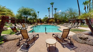 A pool area with lounge chairs and a table, surrounded by palm trees and a clear blue sky.
