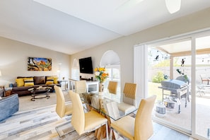 A modern living room with a glass dining table, comfortable seating, a sofa with cushions, a flat-screen TV, and a large window offering a view of the outdoors. - WeeCasa | Dog-friendly house in Chandler with private heated pool, hot tub, AC (Chandler)