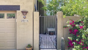 Une maison de couleur beige avec une porte de garage, un portail en métal, un pot de fleurs, un cactus et une bougainvillée rose.